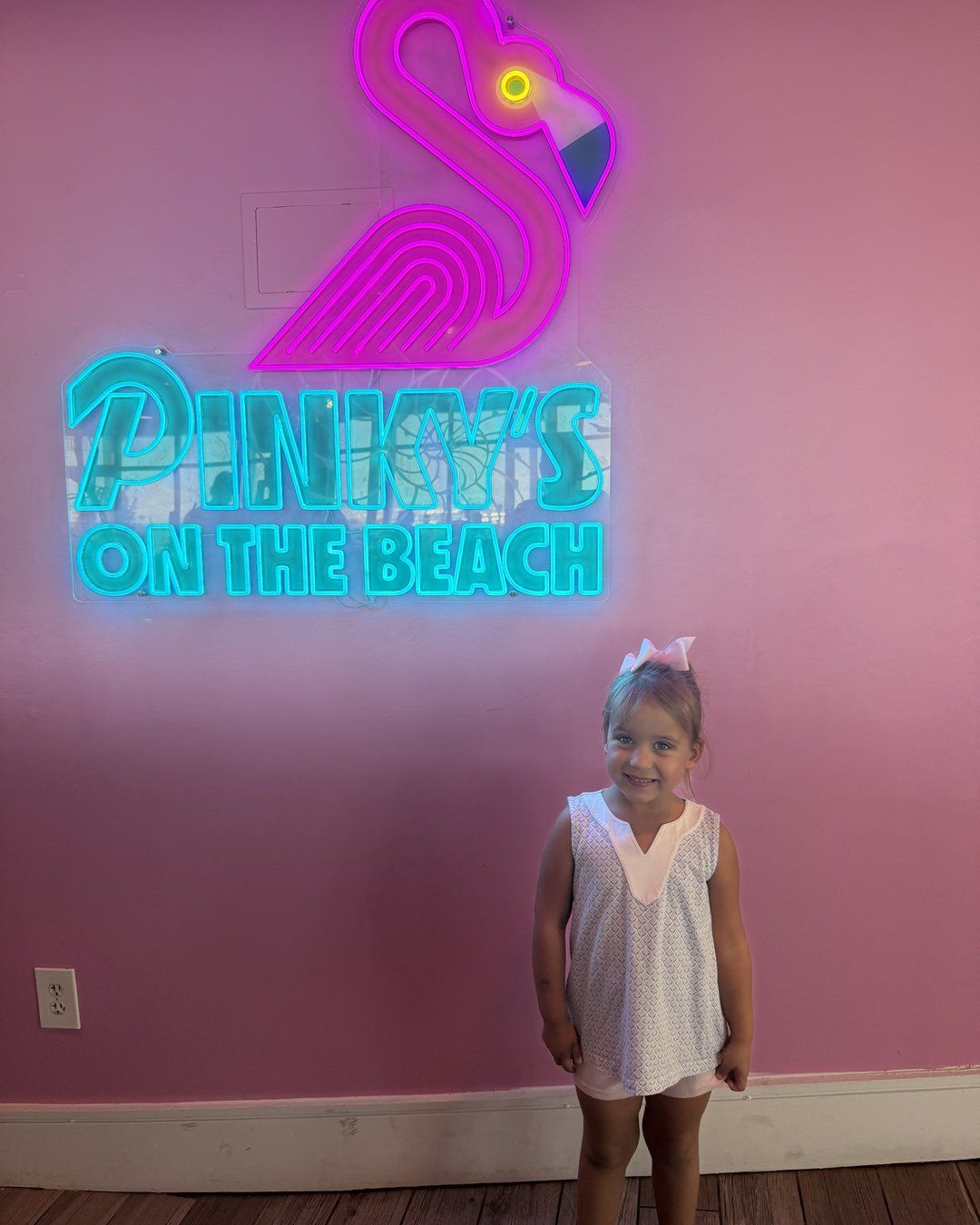 Little girl standing under the neon flamingo Pinky's on the Beach sign on a pink wall