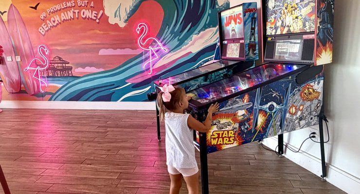 Little girl playing a pinball machine in front of a surf mural at Pinky's on the Beach