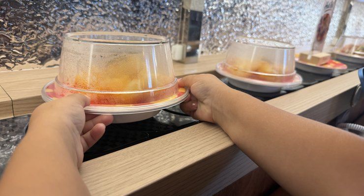 Child's hands lifting a covered plate off the conveyor belt at Revolving Sushi Kingdom