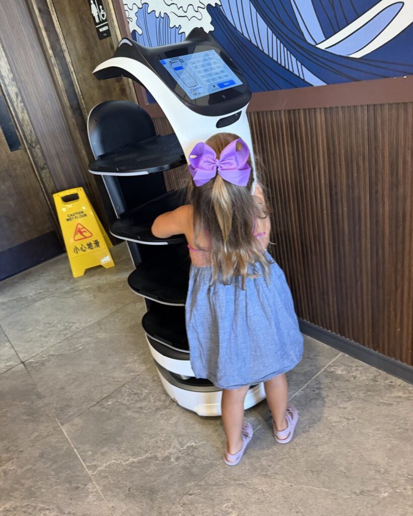 Little girl with a purple bow standing next to a robot server at Revolving Sushi Kingdom